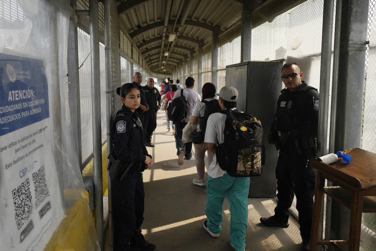 group of migrants walk across a tunnel bridge as officers look on May 2023