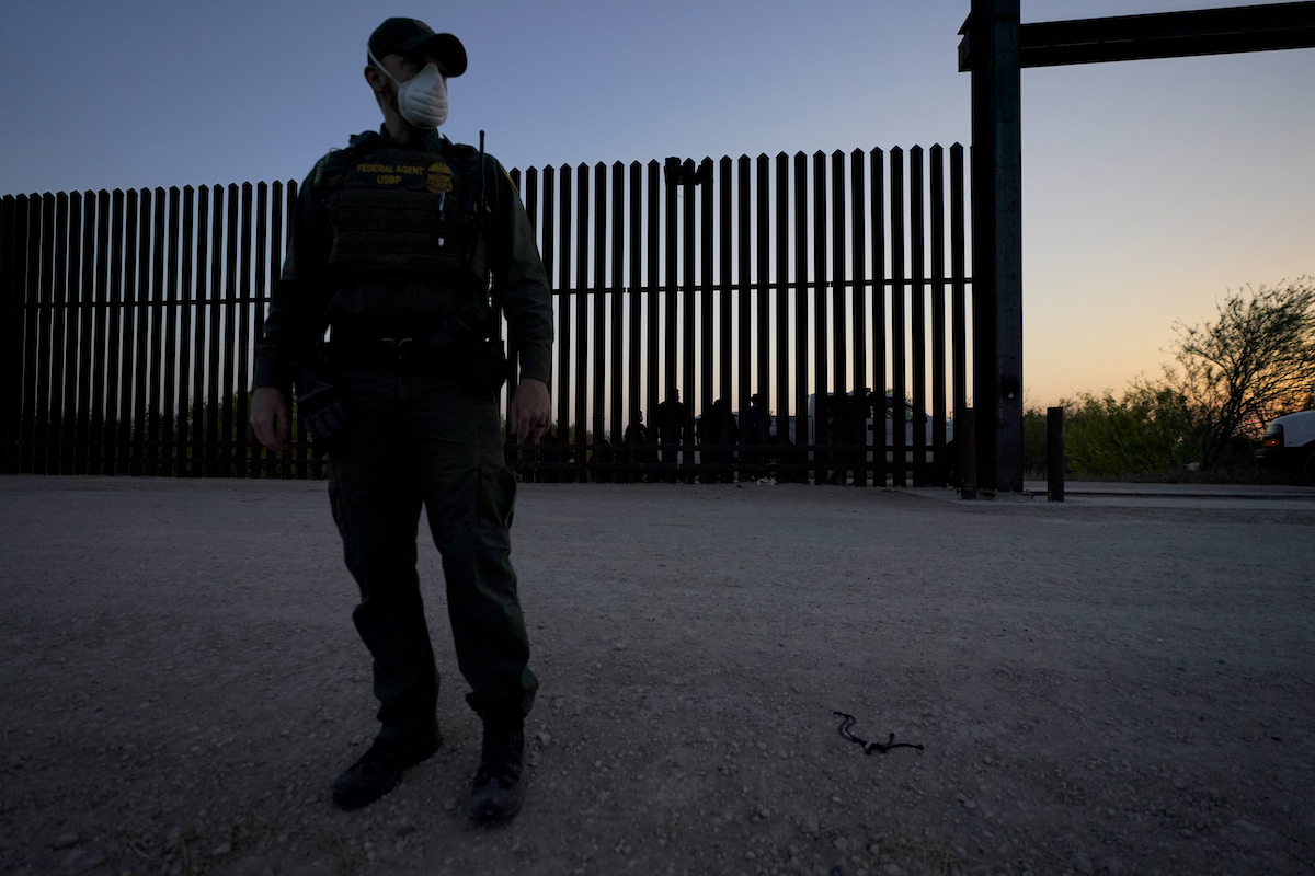 CBP officer looks to the side with border wall behind him