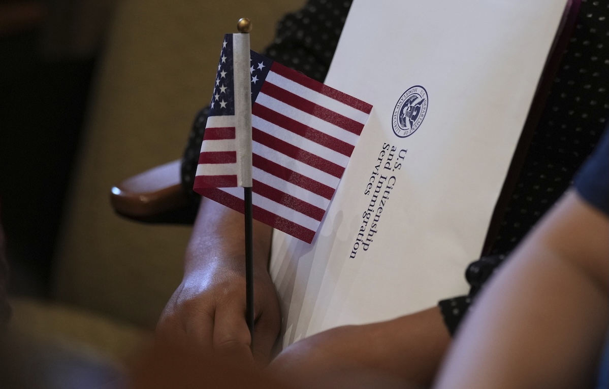 Photo of naturalization ceremony, hand holding US flag + USCIS folder 07-3-2025