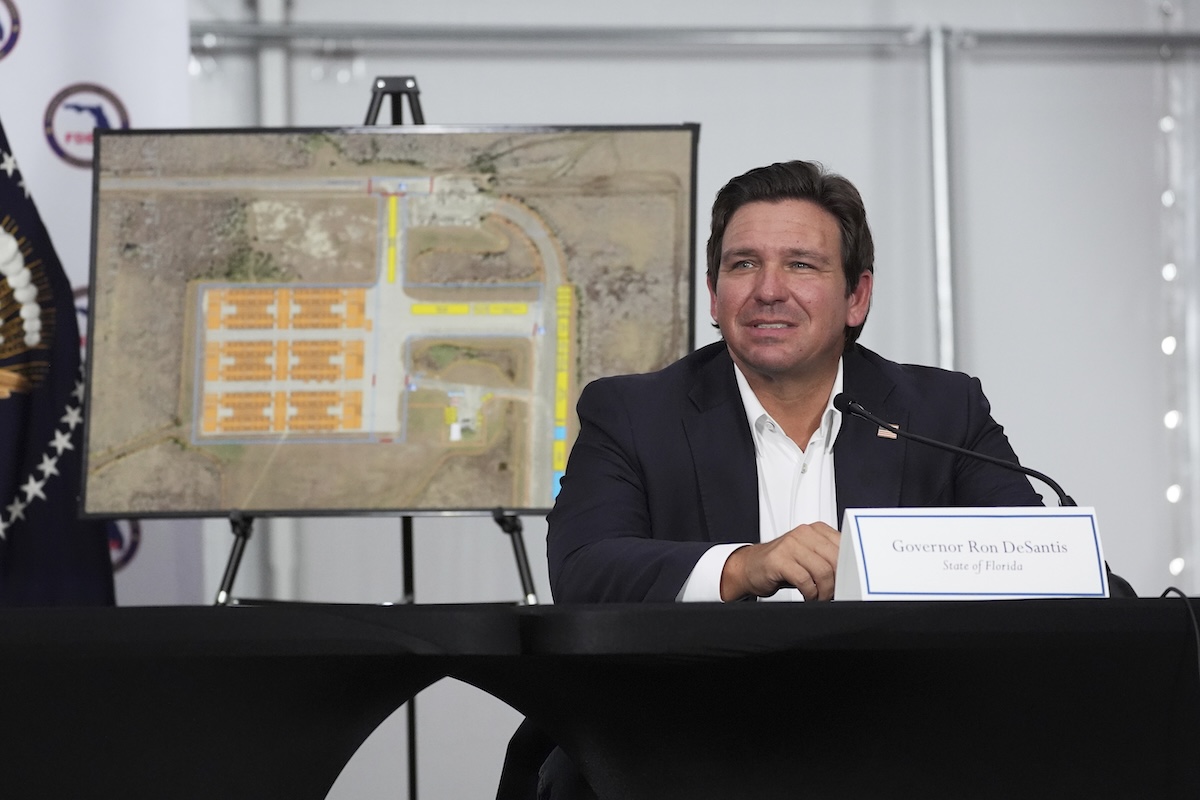 DeSantis with Alligator Alcatraz plans in poster behind him 07-01-2025